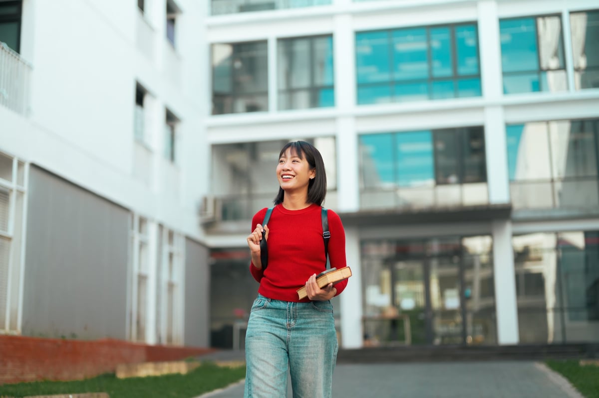 Happy Asian female student walking outdoors on university campus, smiling, carrying books and wearing a backpack