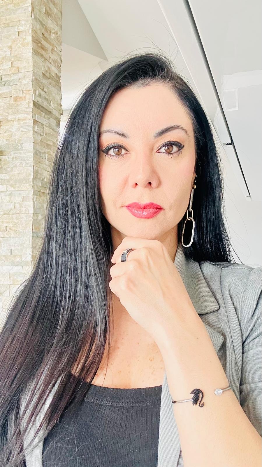 Woman with long black hair wearing pink lipstick and gray blazer, hand near face, posing indoors